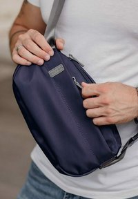 Námořnická crossbody taška vyrobená z hladké tkaniny, s uzávěrem na zip a šedým nastavitelným popruhem. Připevněná dekorativní značka.
