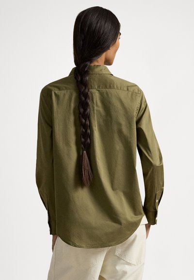 Hosszú, sötét, fonott hajú nő olívazöld gombos ingben és világos bézs nadrágban, háttal állva, fehér háttér előtt.