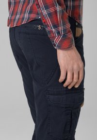 Navy cargo pants feature multiple pockets and textured fabric. Paired with a red and white checkered shirt, showing button details.