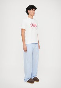 T-shirt branca de manga curta com gráfico vermelho de "AMOR"; combinado com calças largas listradas verticais em azul claro; sapatos castanhos de slip-on.