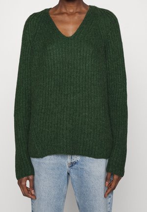 Pull en maille côtelée vert foncé avec manches longues et col en V porté avec un jean en denim bleu clair.