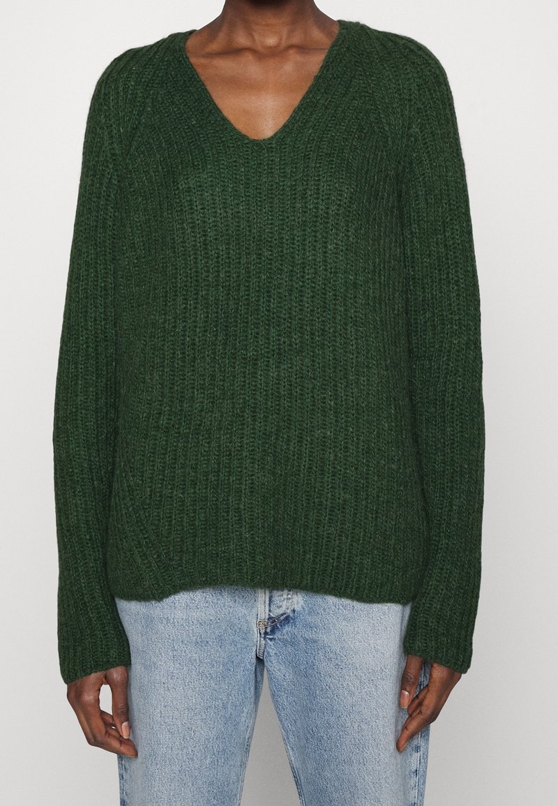 Pull en maille côtelée vert foncé avec manches longues et col en V porté avec un jean en denim bleu clair.