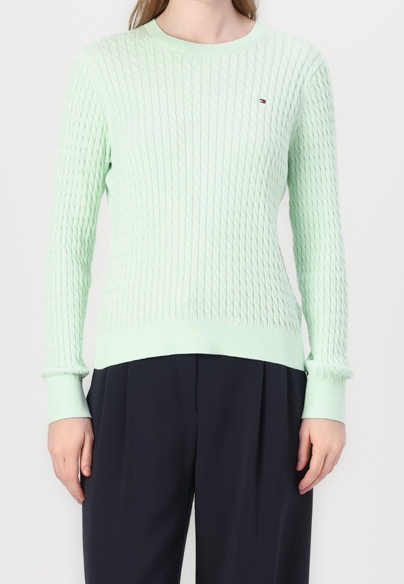 Femme portant un pull à manches longues en maille torsadée vert clair associé à un pantalon noir ample sur un fond uni.