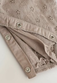 Hellbrauner Stoff mit gestickten Blumen, versehen mit metallenen Druckknöpfen und gerafftem elastischen Detail, gezeigt in einer Nahaufnahme.