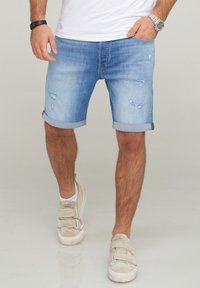 Pantalones cortos de mezclilla azul claro con dobladillos enrollados, que presentan bordes deshilachados y detalles desgastados. Combinados con zapatillas beige y una camisa blanca.