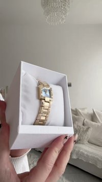 Orologio in metallo dorato con quadrante quadrato, quadrante azzurro chiaro e bracciale a maglie, esposto in una scatola bianca con un inserto morbido.