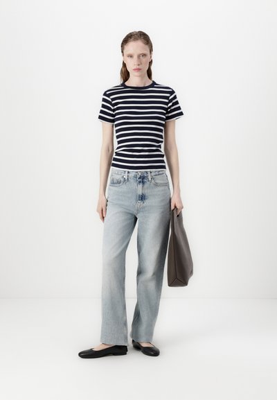 T-shirt à manches courtes rayé navy et blanc, jean large bleu clair, chaussures plates noires et sac fourre-tout gris sur fond blanc.