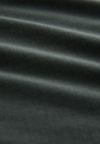 Temno zelena tkanina z mehko teksturo, ki ima nežne valove in gladko površino, kar poudarja njeno lahko in prilagodljivo naravo.