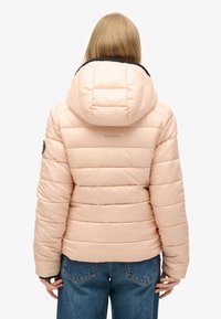 Giacca a vento rosa chiaro con cappuccio regolabile, pannelli cuciti e dettaglio del logo sul retro. Abbinata a jeans blu a vita alta.