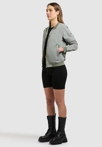 Femme debout portant une veste bomber gris clair, un short noir et des bottes de combat à lacets noires sur un fond clair uni.
