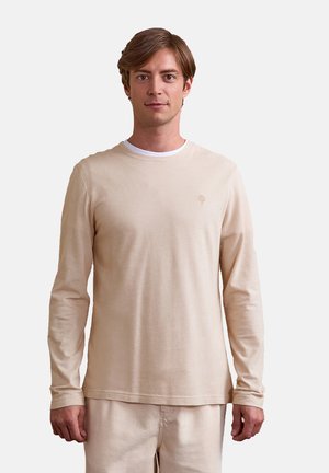 Junger Mann mit hellbraunen Haaren trägt ein beigefarbenes Langarmhemd und passende Hosen, steht vor einem schlichten hellgrauen Hintergrund.