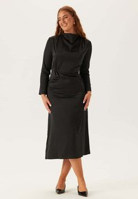 Abito nero a maniche lunghe con scollatura a serpentina, vita aderente e gonna fluida. Tessuto liscio con texture minima, abbinato a tacchi neri.