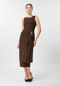 Femme portant une robe midi marron sans manches avec drapé latéral et boucle dorée décorative, chaussée de talons noirs et portant des boucles d'oreilles créoles.