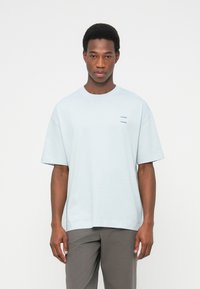 Ανοιχτό μπλε βαμβακερό T-shirt με στρογγυλή λαιμόκοψη και κοντά μανίκια, με μικρό τυπωμένο λογότυπο στο στήθος. Συνδυάζεται με γκρι παντελόνι.