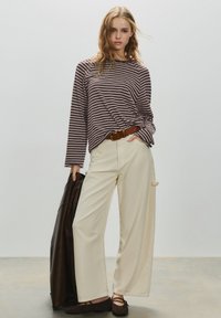 Ung kvinde iført stribet langærmet skjorte, cremefarvede bukser med vide ben, brun bælte og flade sko, som holder en mørk jakke mod en ensfarvet baggrund.