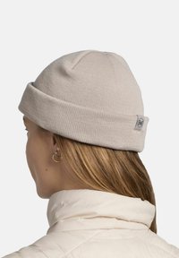 Light beige beanie in a ribbed knit texture with a folded cuff. Features a small grey logo tag on the side. Smooth and fitted design.