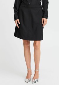 Schwarzer Rock mit einem Bund, gerader Schnitt, knielang, aus glattem Stoff. Kombiniert mit glänzenden silbernen High-Heels.