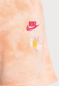 Camiseta de Nike en melocotón claro con un patrón de tie-dye, que presenta un logo rojo y una margarita bordada con centro amarillo y pétalos blancos.