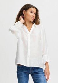 White button-up shirt with a V-neck, loose fit, and long sleeves featuring gathered cuffs. Textured fabric adds subtle detail. Paired with blue jeans.