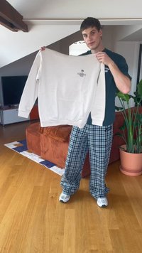 Vīrietis stāv viesistabā, turot baltu sporta krekls ar mazu "ATHENS" logotipu, ģērbies melnās rūtainās biksēs un baltos sporta apavos.