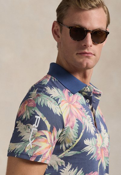 Άνδρας που φορά γυαλιά ηλίου με καφέ σκελετό τύπου χελώνας και μπλε πόλο πουκάμισο με ροζ και πράσινα τροπικά φύλλα, καθώς και λευκό λογότυπο στο μανίκι.