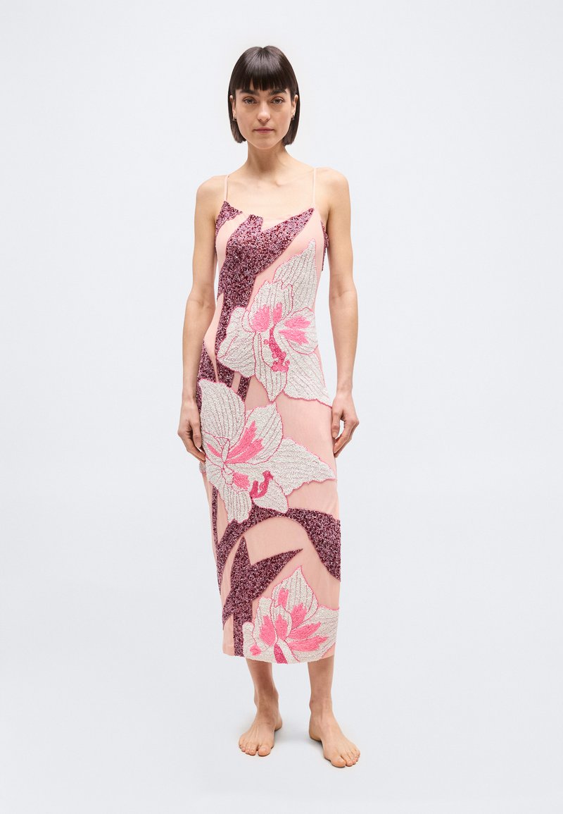 Μια γυναίκα στέκεται ξυπόλητη φορώντας ένα ροζ midi φόρεμα χωρίς μανίκια με μεγάλα λουλουδάτα και αφηρημένα σχέδια από παγιέτες, πάνω σε απλό φωτεινό φόντο.