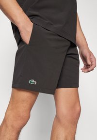 Sorte shorts laget av lett materiale, med en sidelomme og en grønn Lacoste krokodille-logo. Enkelt design, med svingte kanter.
