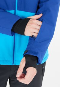 Giacca blu e turchese con una texture liscia, polsino con zip e chiusura in Velcro. La manica interna nera si estende sotto lo strato esterno.