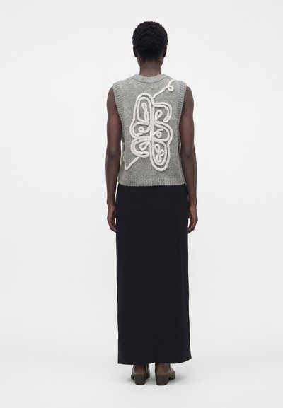 Personne portant un gilet en tricot gris sans manches avec un motif abstrait en forme de corde blanche dans le dos, associé à une longue jupe noire et des talons, debout de dos.