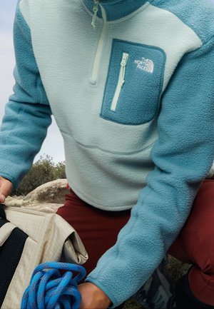 Person trägt eine türkise und weiße Fleecejacke mit einer Reißverschlusstasche auf der Brust, hält im Freien ein blaues Kletterseil und einen beigen Rucksack.