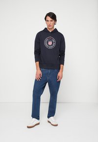 O moletom azul marinho com capuz apresenta uma ilustração circular com detalhes em vermelho, branco e azul. Combinado com jeans azuis largos e ténis brancos.