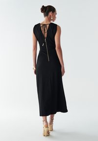 Melns midi kleita ar pieguļu pie vidukļa, atvērtu muguras dizainu un zelta rāvējslēdzēja detaļu. Teksturēts audums un piedurkņu trūkums ar savāktiem pleciem.