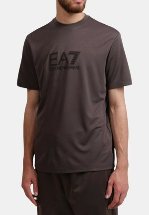 Uomo che indossa una t-shirt a maniche corte EA7 Emporio Armani marrone scuro e pantaloni abbinati, in piedi contro uno sfondo chiaro e uniforme.
