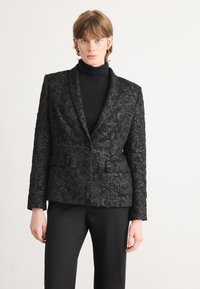 Zwarte gebreide blazer met bloemenpatroon, slim fit, pieklapels, twee voorkruiszakken, gedragen over een zwarte turtleneck, gecombineerd met zwarte broek.