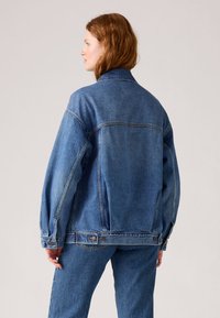 Modra denim jakna z udobnim krojem, dolgimi rokavi, ožjim ovratnikom in gumbi. Poudarjena z kontrastnimi šivi in subtilnim bledenjem.