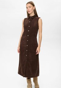 Ženska stoji oblečena v brezrokavni rjav gumbasti midi obleki iz kordovih in svetlo rjavih semiš škornjih, v ozadju pa je enostavna siva podlaga.