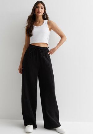 Top corto blanco combinado con pantalones negros de pierna ancha. Los pantalones presentan una tela texturizada, con cintura de cordón y bolsillos laterales.