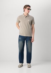 Beige polo skjorte med en liten logo, kombinert med mørkeblå jeans og grønne solbriller. Outfiten har korte ermer og klassisk krage.