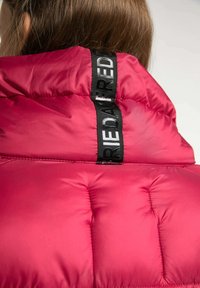 Röd puffjacka med en kviltad design med fyrkantiga sektioner och en svart logotypetikett på kragen. Slät, blank yta.
