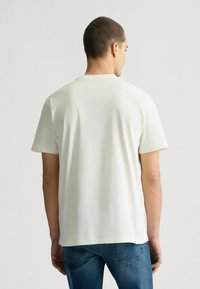T-shirt a maniche corte e collo rotondo in tessuto avorio, caratterizzato da una vestibilità comoda e una superficie liscia. La vista dal retro mostra un design semplice senza decorazioni.