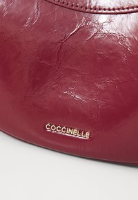 Burgundy leather handbag with a textured finish, featuring a gold "Coccinelle" logo at the bottom. Smooth, curved design.
