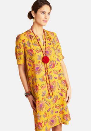 Frau trägt ein gelbes Kleid mit bunten Blumenmustern, eine rote Perlenkette mit rotem Bommel, goldene Ohrringe und ein mehrfarbiges Armband.