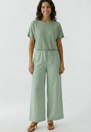 Lächelnde Frau mit lockigem Haar, die ein hellgrünes Kurzarmoberteil und passende weit geschnittene Hosen mit Bindegürtel in der Taille trägt, stehend in braunen Sandalen.