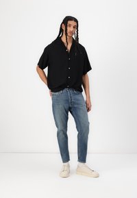 Schwarzes Kurzarm-Hemd mit Knöpfen, hellblaue Jeans und beigefarbene Sneaker. Das Hemd hat eine lockere Passform; die Jeans sind an den Knöcheln schmal zulaufend.
