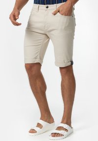 Beżowe bawełniane szorty z podwiniętymi mankietami, z zapięciem na guzik, przednimi kieszeniami oraz kontrastowym akcentem na mankietach. Noszone z białymi sandałami.
