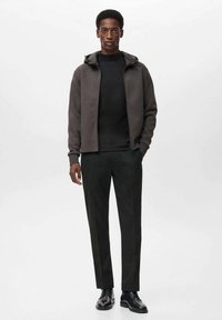 Mladic u sivoj dukserici s patentnim zatvaračem, crnom rolkom, crnim hlačama i crnim kožnim cipelama s vezicama stoji protiv jednostavne pozadine.