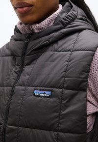 Κοντινό πλάνο ενός ατόμου που φοράει μαύρο καπιτονέ γιλέκο Patagonia πάνω από ένα ροζ υφή πουλόβερ, με το λογότυπο της μάρκας στο γιλέκο.