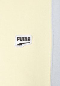 Naszywka z logo PUMA na jasnożółtej tkaninie, otoczona miękką jasnoszarą ramk�ą. Faktura prążkowanego materiału o czystym, nowoczesnym designie.