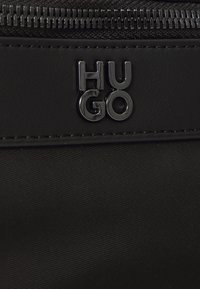 Melnas auduma soma ar gludu faktūru, izteiktu rāvējslēdzēju un izcilu melnu metāla logotipu "HUGO". Vienkāršs, moderns dizains.