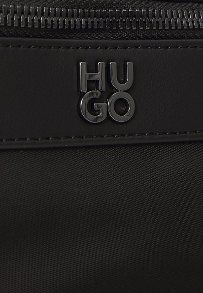 Sac en tissu noir avec une texture lisse, une fermeture éclair proéminente et un logo noir en métal "HUGO" en relief. Design simple et moderne.